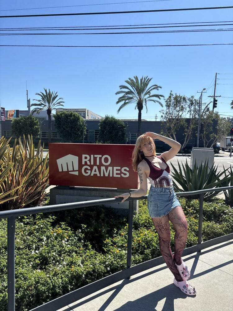 Steph posando ao lado da placa da Riot Games, editada pra dizer Rito Games